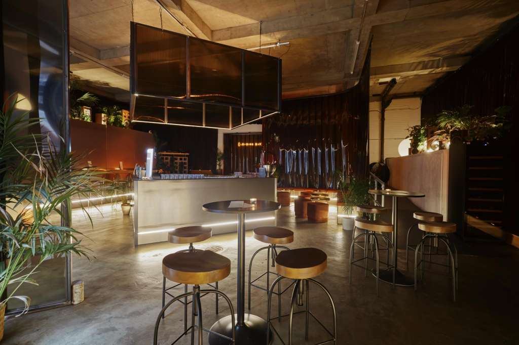 Industrial event space with bar counter, high seating, ambient lighting, and potted plants throughout.