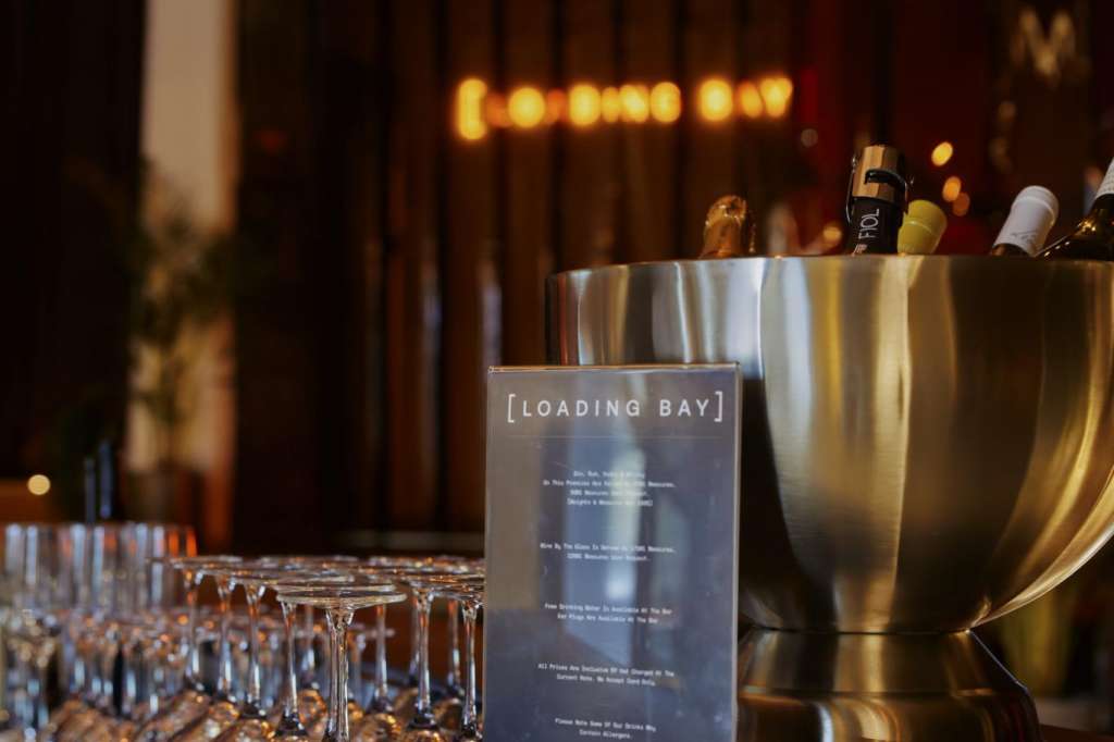 Bar setup with champagne bucket, glasses, and Loading Bay signage on dark counter with ambient lighting.