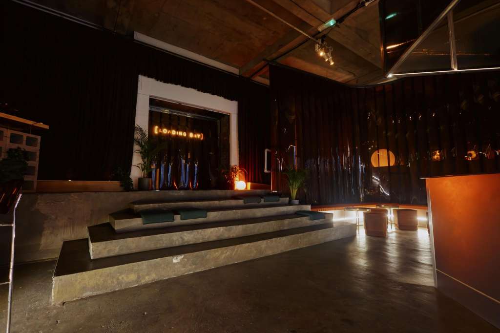 Industrial event space with concrete tiered seating, stage with neon signage, warm ambient lighting, and decorative plants.