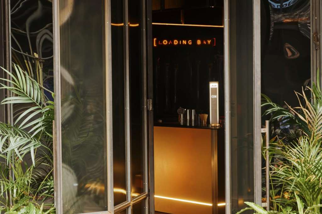 Loading Bay entrance with glass doors, neon signage, potted plants, and modern interior lighting visible through open doors.