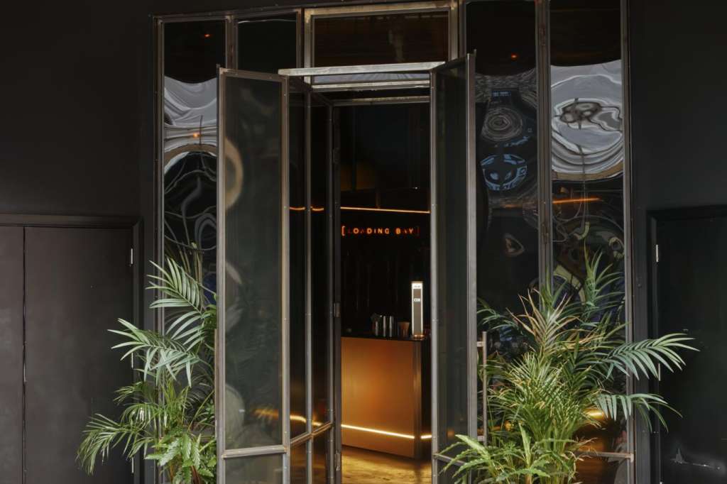 Exterior storefront entrance with Loading Bay signage, glass doors, and potted plants flanking the entry.