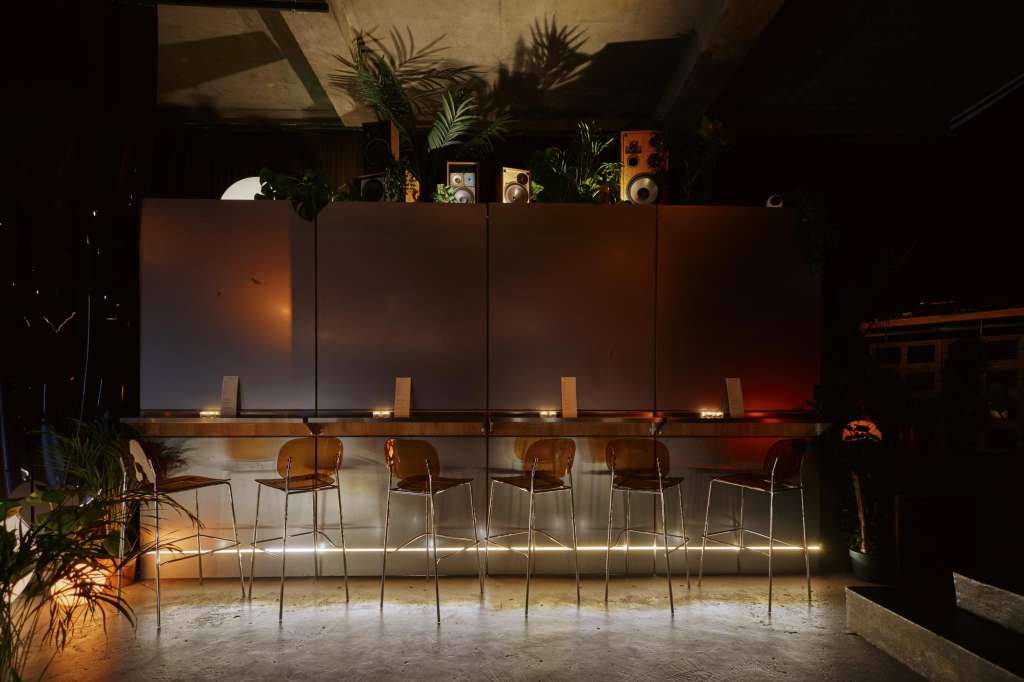 Industrial space with bar seating, modern chairs, potted plants, and warm ambient lighting against a dark backdrop.