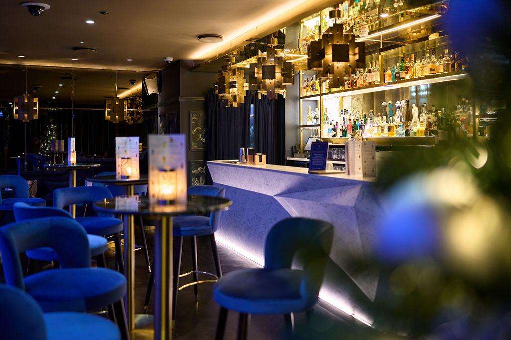Bar area with blue seating, illuminated counter, and backlit shelving displaying spirits bottles.