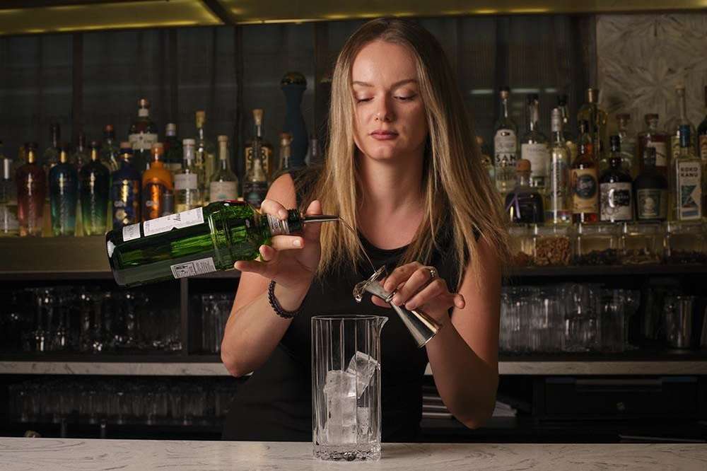 Bartender pouring wine at a bar counter with illuminated bottle display behind.