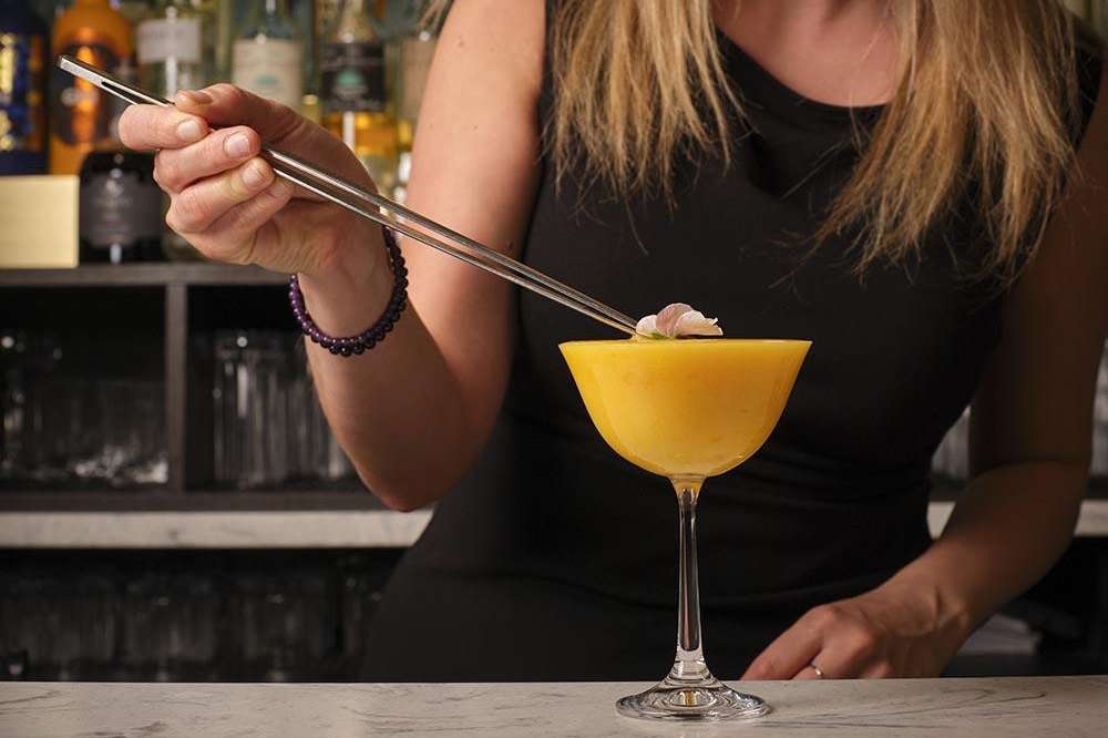 Bartender preparing a yellow cocktail with tweezers at a bar counter with bottles in background.