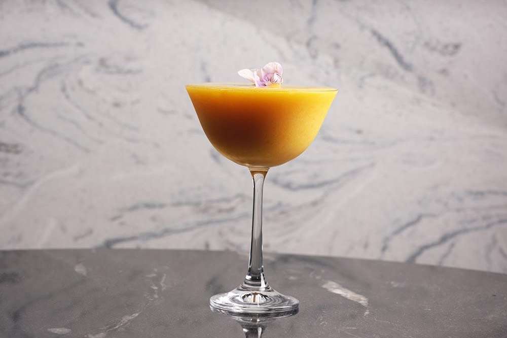 Cocktail with golden liquid and pink flower garnish on marble surface against patterned wall.