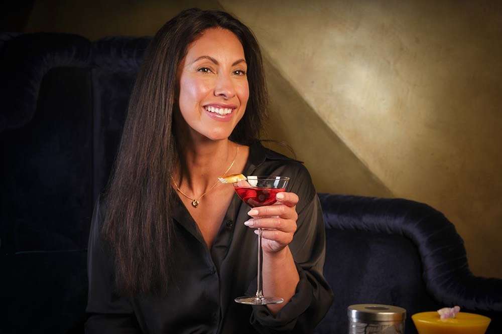 Woman in black shirt holding cocktail, seated on dark sofa in lounge bar with warm lighting.