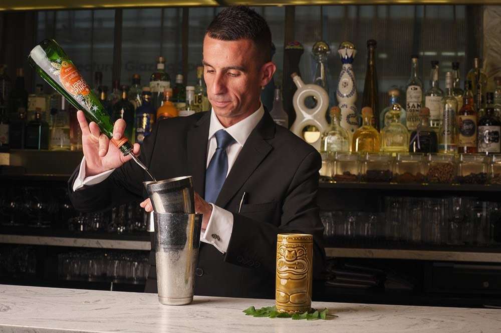 Bartender in formal attire pouring champagne at a marble bar counter with extensive bottle display behind.