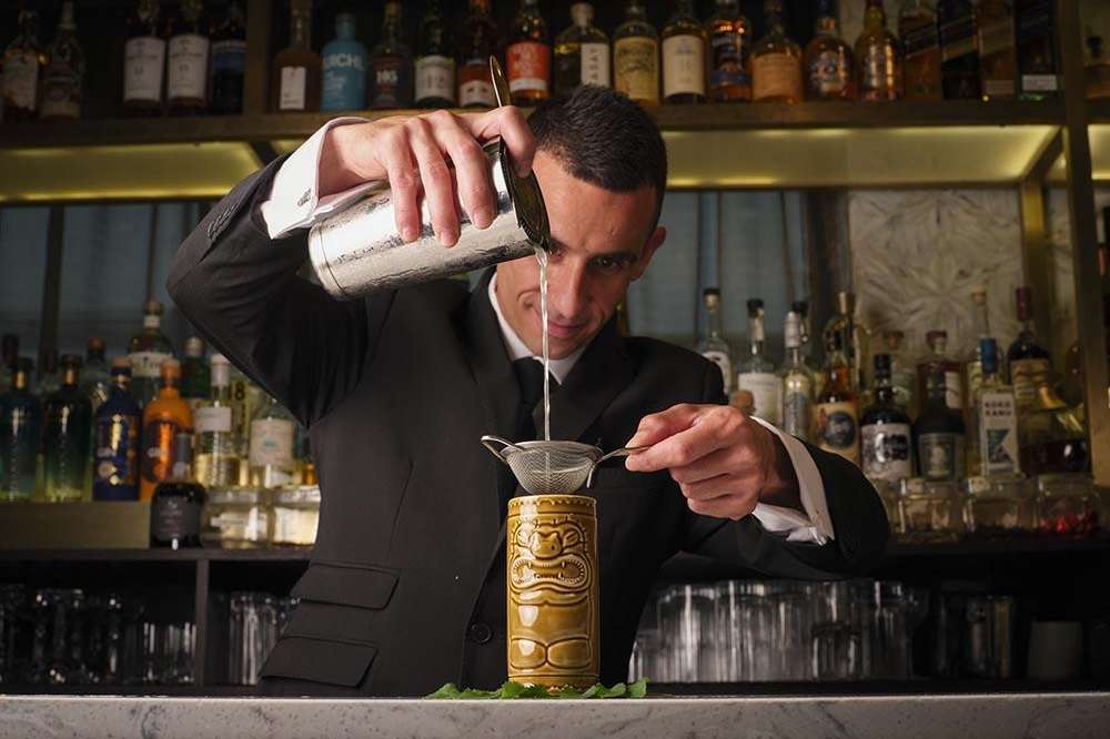 Bartender in black suit pouring cocktail at Lounge Bar with illuminated bottle display behind.