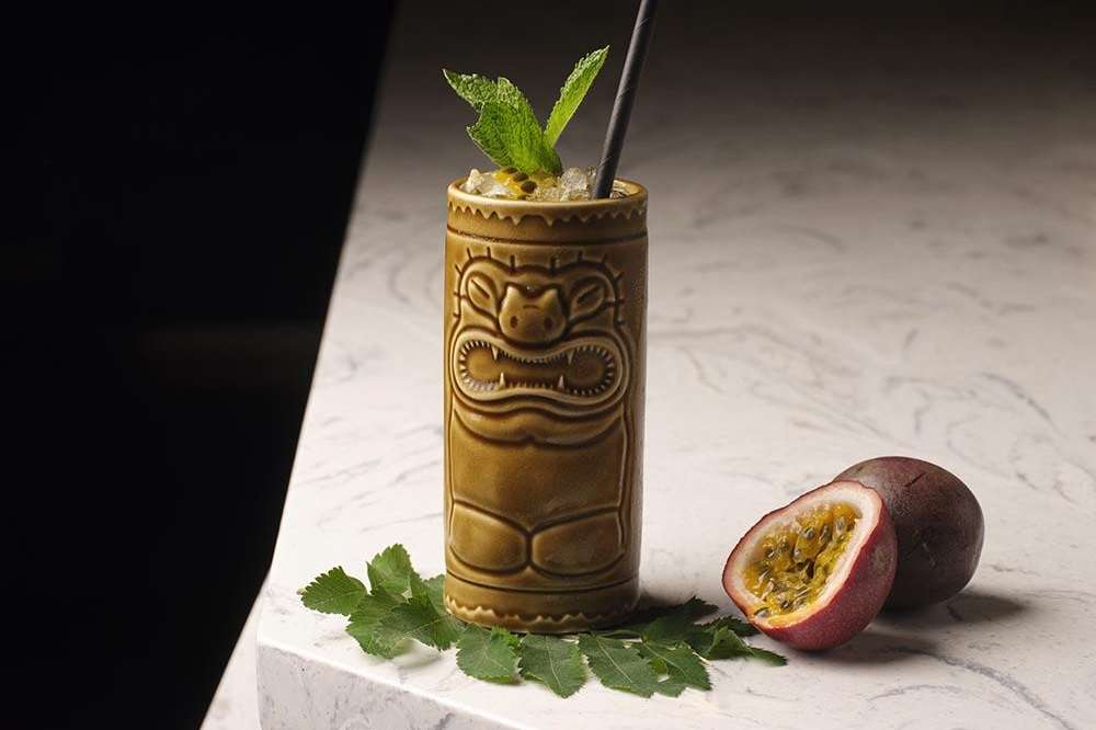 Tropical cocktail in ornate gold tiki mug with mint and black straw, fresh passion fruit beside on marble surface.