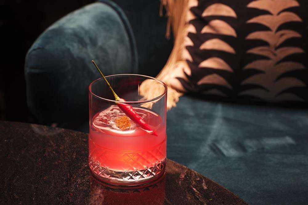 Craft cocktail with red liquid and garnish on dark bar counter with decorative backdrop.