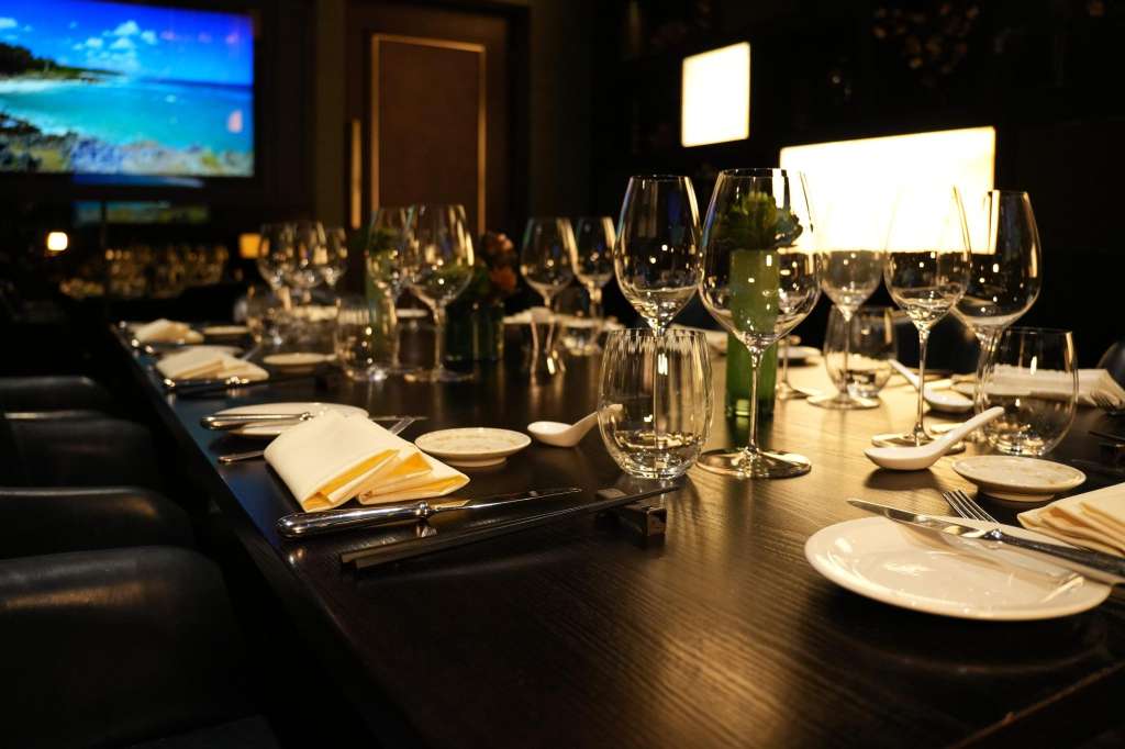 Long dining table set with wine glasses, place settings, napkins, and formal tableware under warm lighting.