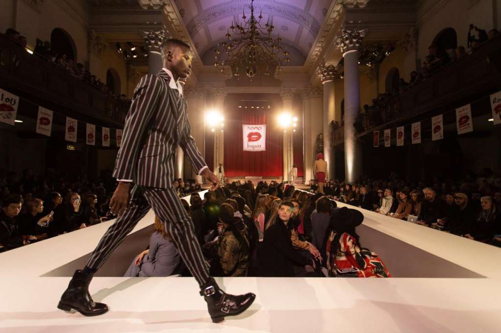 Fashion show runway with model in striped suit, seated audience on both sides, ornate ceiling with chandelier and stage backdrop.