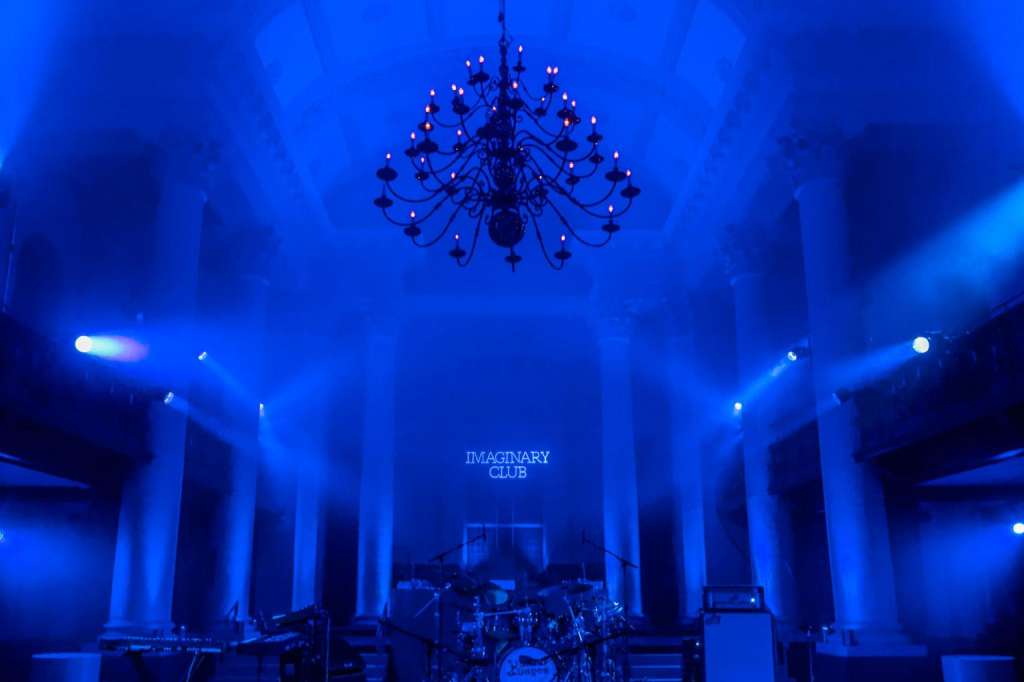 Grand hall with blue uplighting, chandelier, stage setup with drum kit and sound equipment.
