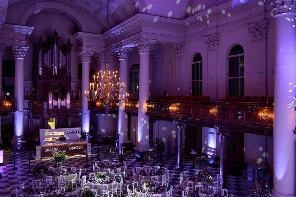 Grand hall with round tables, chiavari chairs, and purple uplighting. Ornate ceiling, chandelier, and organ visible.