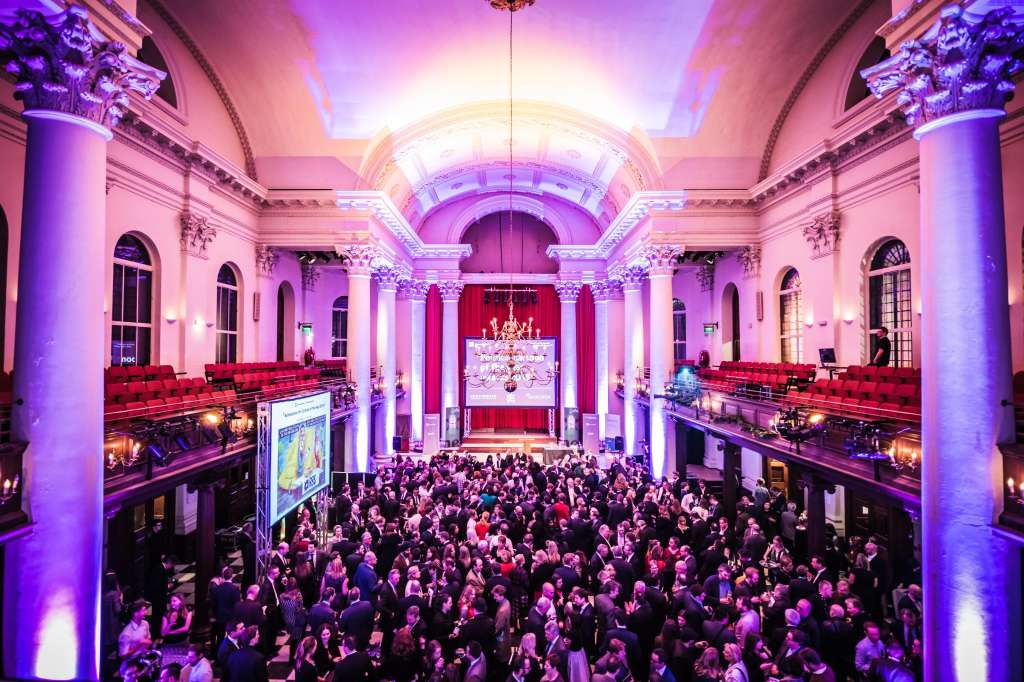 Grand hall with ornate columns and arched ceiling, lit in purple and pink, filled with standing guests and a stage with red curtains.