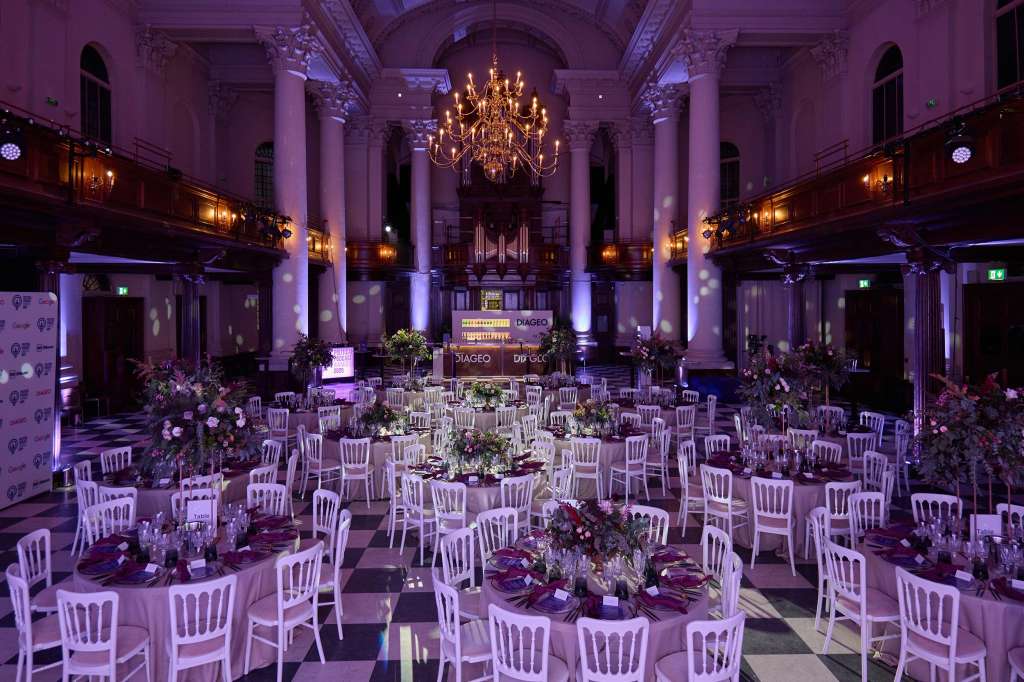 Grand hall with round tables, white chairs, floral centerpieces, and purple ambient lighting under ornate ceiling.