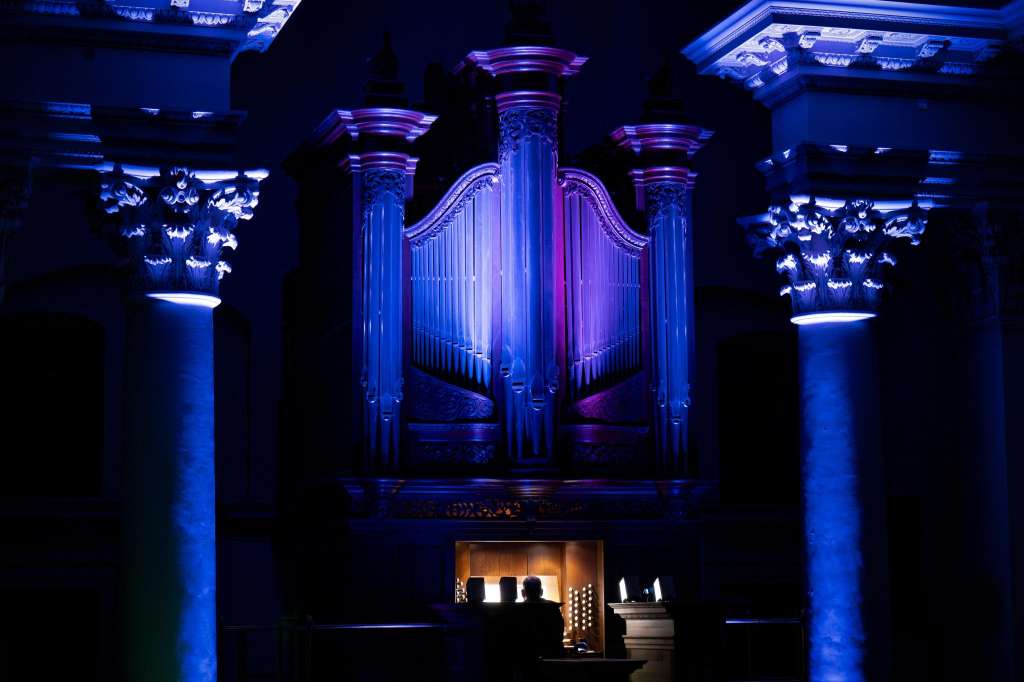 Grand pipe organ illuminated with blue and purple lighting in a classical hall with ornate columns and capitals.
