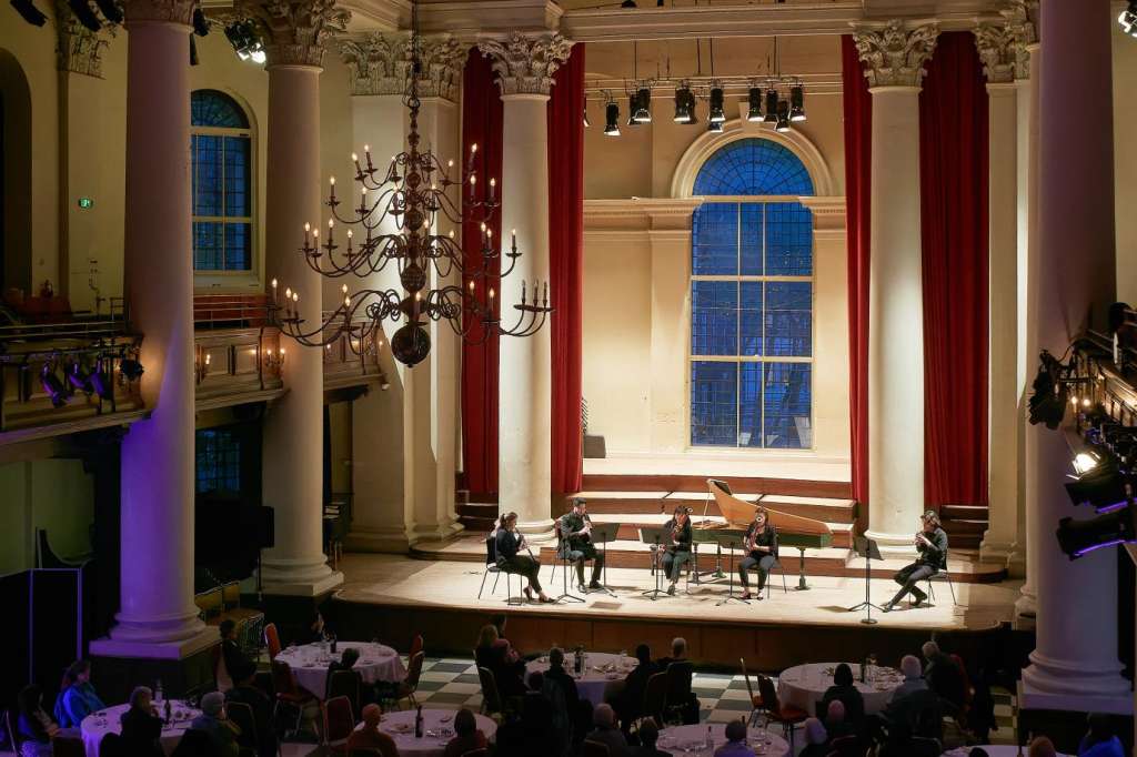 Grand hall with ornate ceiling, columns, and stage set for seated dinner with round tables and purple lighting.