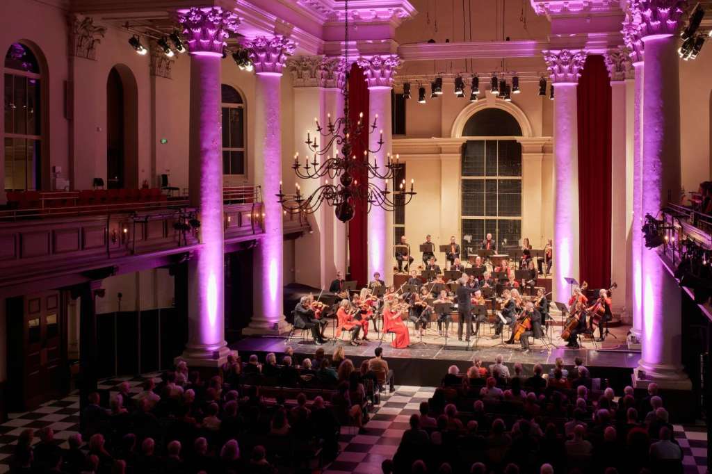 Grand hall with vaulted ceiling, purple uplighting, ornate columns, and seated audience facing a stage with orchestra.