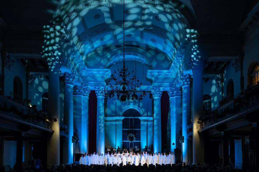 Grand hall with blue architectural projection mapping, chandelier, and performers in white robes on stage.
