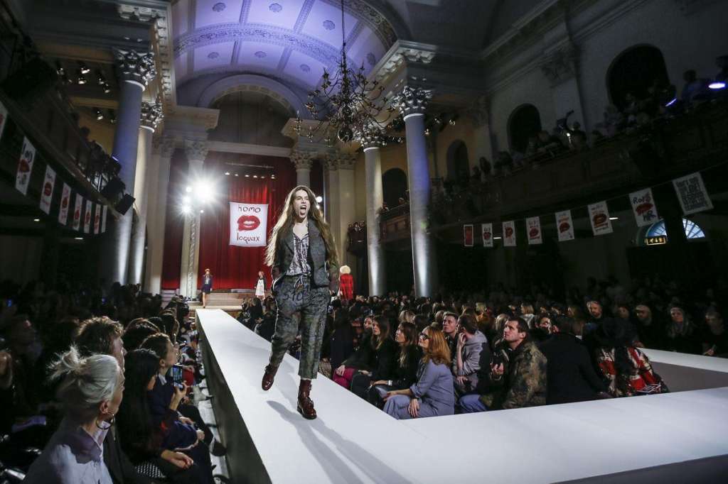 Fashion show runway with model walking down white catwalk, seated audience on both sides, ornate hall with columns and chandelier.
