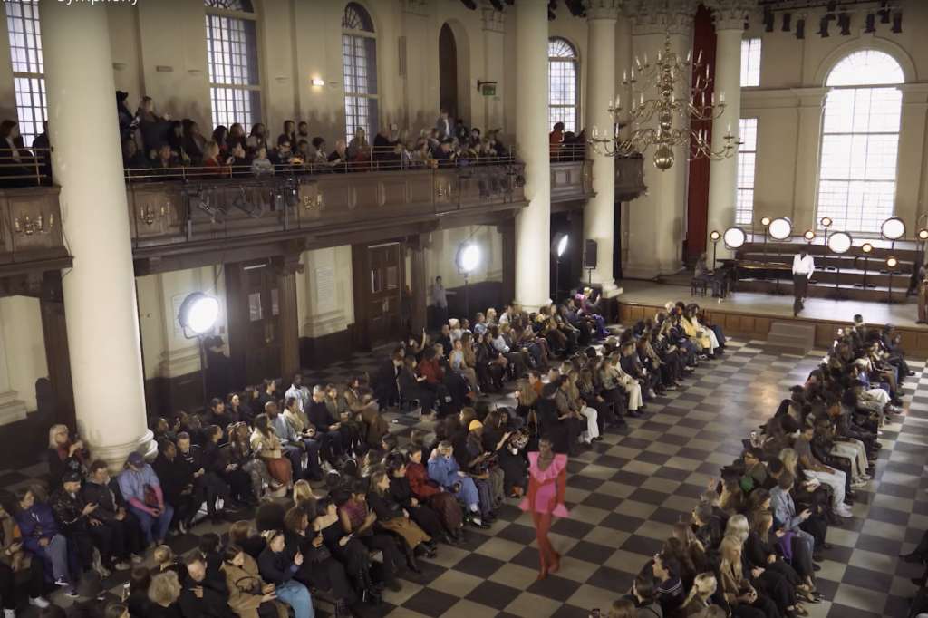 Grand hall with high ceilings, columns, and gallery seating. Large crowd gathered for a standing event with central stage area.
