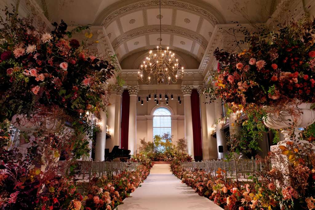 Grand hall with ornate ceiling, chandelier, and elaborate floral installations lining a central aisle.