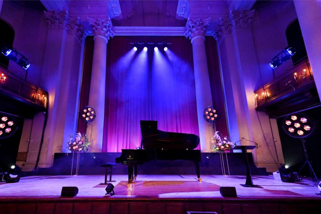 Grand hall stage with grand piano, ornate columns, purple uplighting, and floral arrangements.