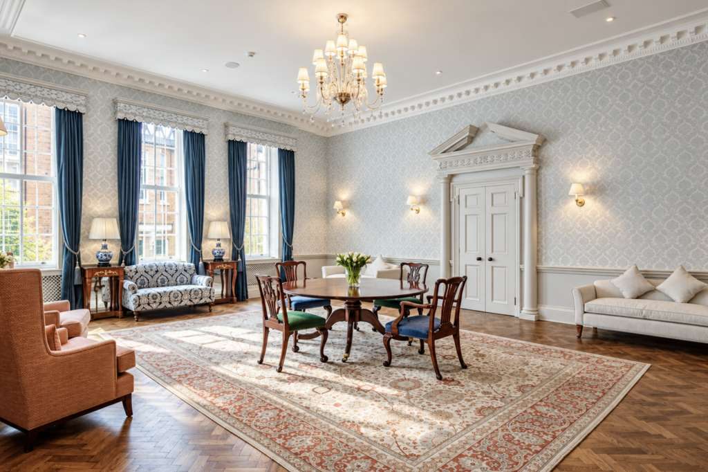Elegant reception room with round dining table, upholstered seating, chandelier, and tall windows with blue drapes.