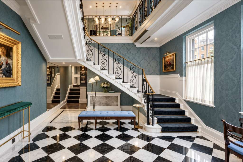 Grand entrance hall with dual staircase, black-and-white marble flooring, blue damask walls, and gold-framed artwork.