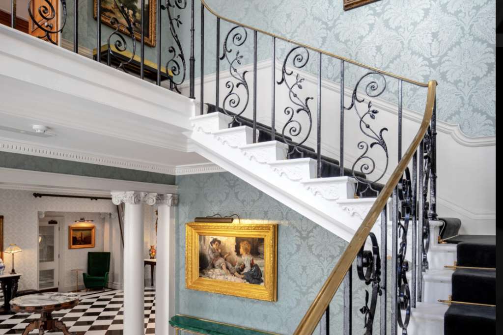 Grand staircase with ornate wrought-iron railings, blue damask wallpaper, gold-framed portraits, and black-and-white checkered floor below.
