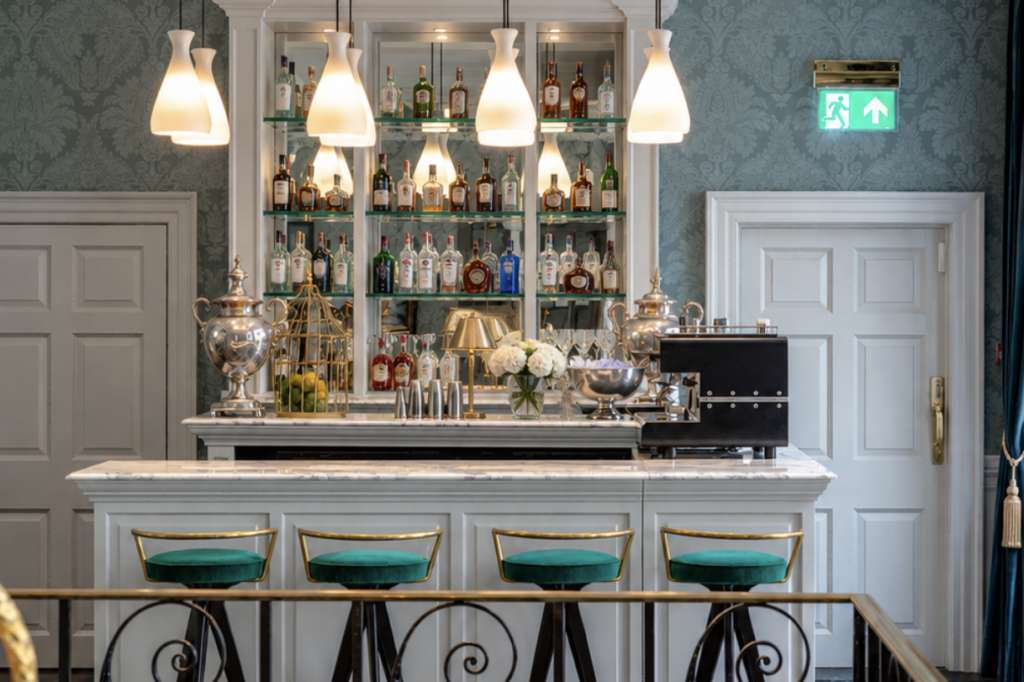 Elegant bar with white counter, turquoise bar stools, pendant lighting, and fully stocked shelving behind.