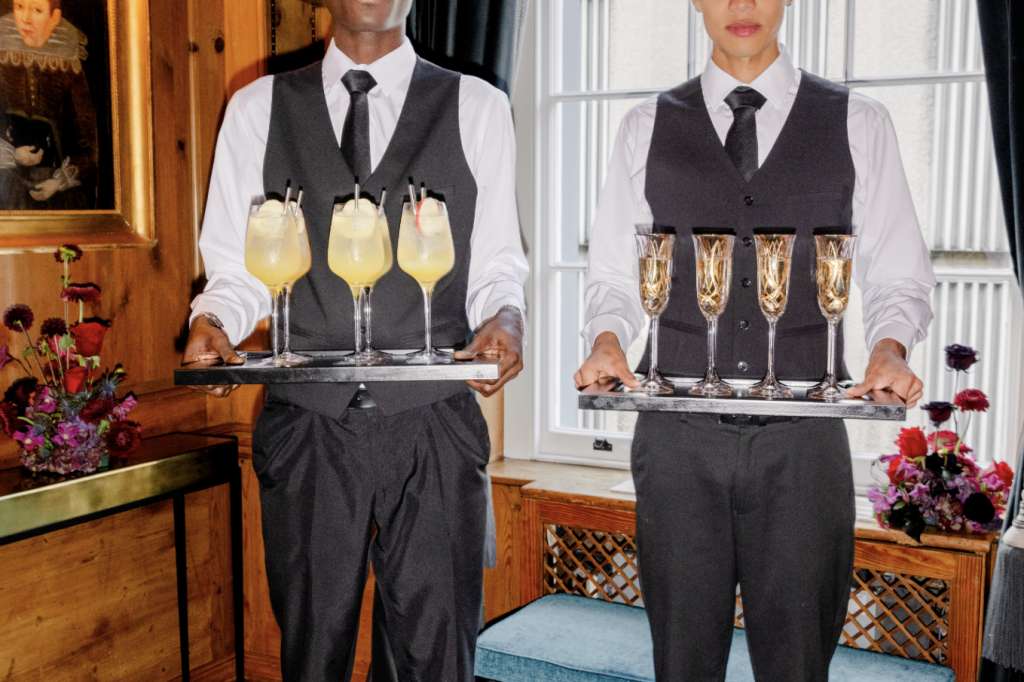 Two staff members in formal waistcoats holding trays of champagne and cocktails in an elegant bar with wood paneling and large windows.