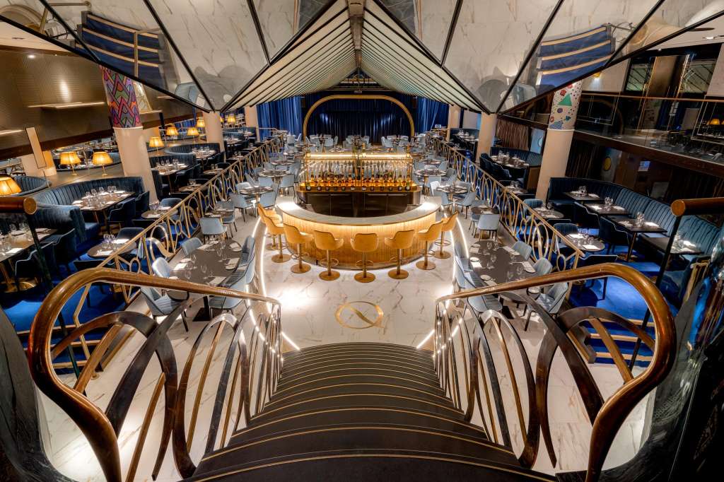 Grand dining room with central bar, round tables set with place settings, blue velvet chairs, and vaulted ceiling with dramatic lighting.