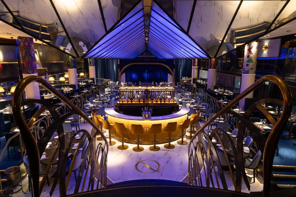 Grand dining room with blue uplighting, central bar, stage, and round tables set for service.