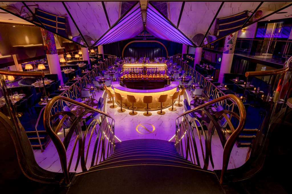 Large event space with purple and gold lighting, central bar, tiered seating, and dramatic vaulted ceiling.