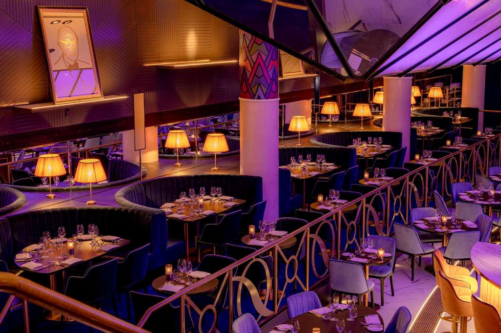 Upscale dining room with banquette seating, round tables set with glassware, warm lighting, and purple ambient lighting throughout.