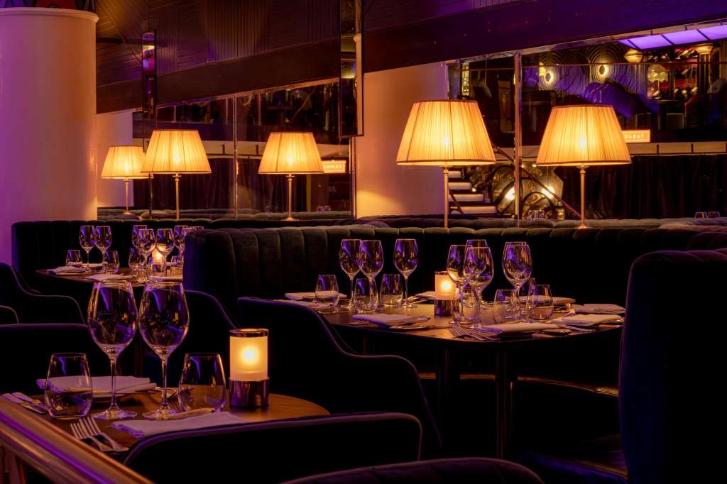 Upscale dining space with black banquettes, set tables with wine glasses, warm table lamps, and purple ambient lighting.