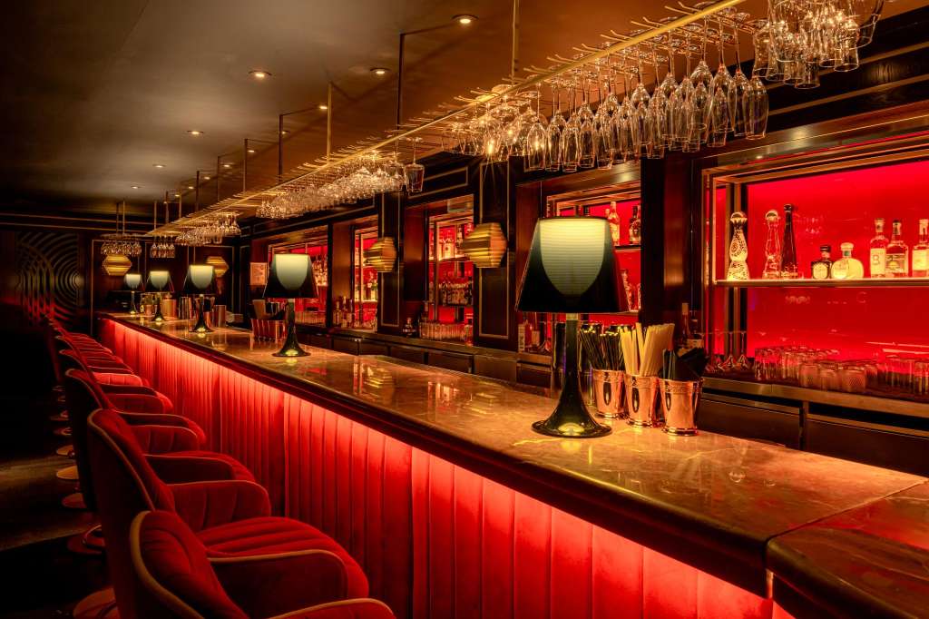 Bar counter with red upholstered seating, hanging glassware, and illuminated spirit bottles on shelves.