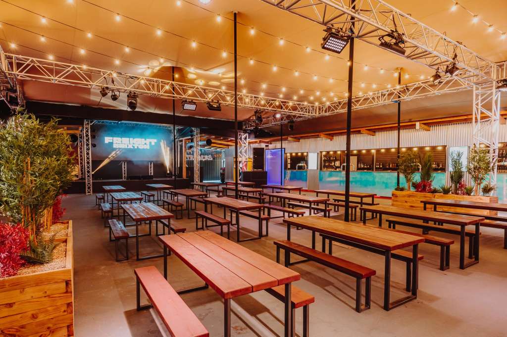 Industrial warehouse space with picnic tables, stage, screens, and warm string lighting. Decorated with potted plants.