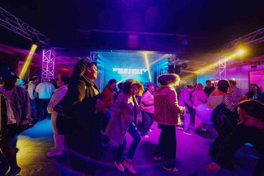Large warehouse space with crowd dancing under vibrant purple and blue stage lighting, projection screen, and overhead trusses.