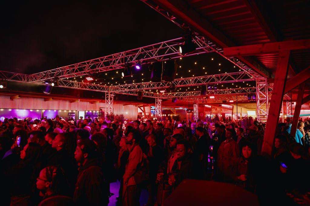 Large warehouse venue with industrial truss ceiling, red and purple ambient lighting, and crowd of guests mingling.
