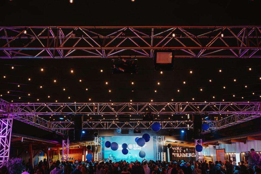 Large warehouse venue with stage, suspended blue balloons, purple truss lighting, and crowd gathered for event.
