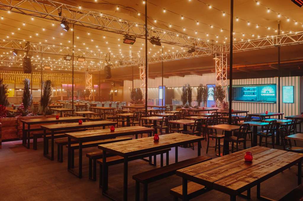 Industrial warehouse space with long wooden tables, benches, string lighting overhead, and decorative pampas grass installations.