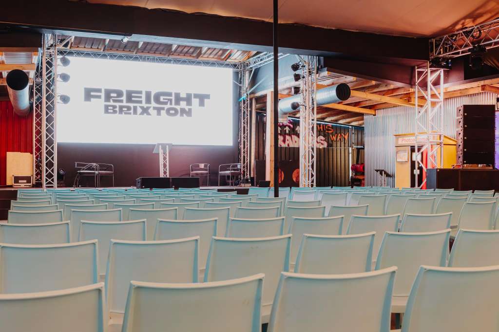 Full venue with rows of light blue chairs facing a stage, large projection screen displaying Freight Brixton branding, and industrial truss lighting.