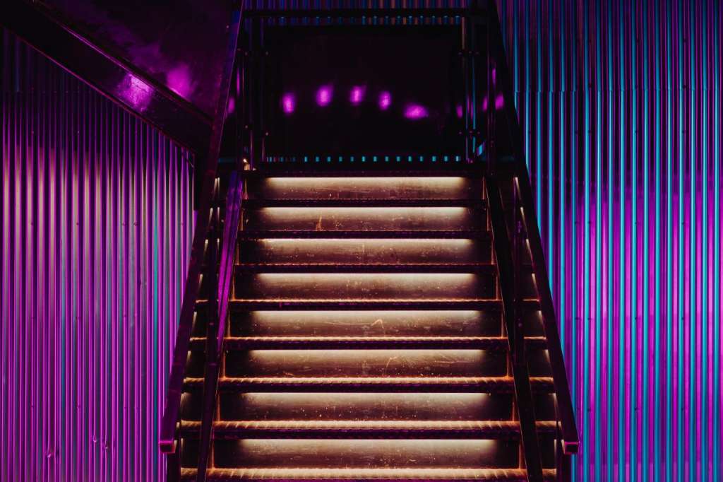 Industrial staircase with warm step lighting, surrounded by vibrant pink and blue neon wall panels and overhead arches.
