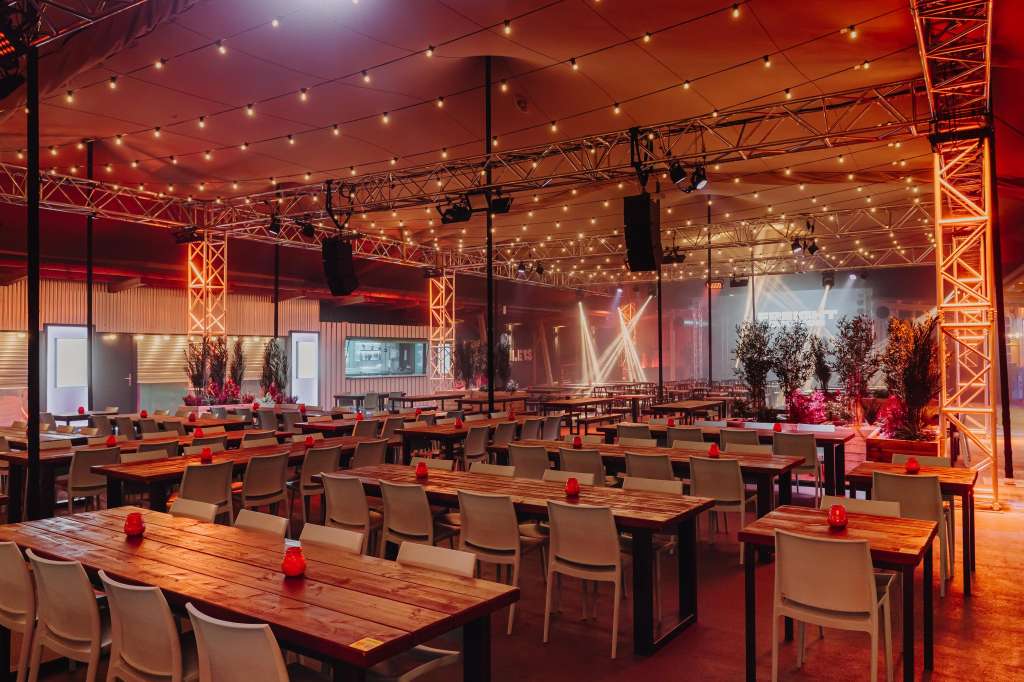 Large industrial venue with long wooden tables, modern chairs, red ambient lighting, exposed steel trusses, and floral installations.