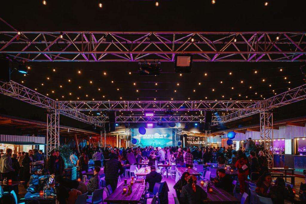 Large industrial venue with exposed truss, purple and blue stage lighting, crowd of guests, and central screen display.