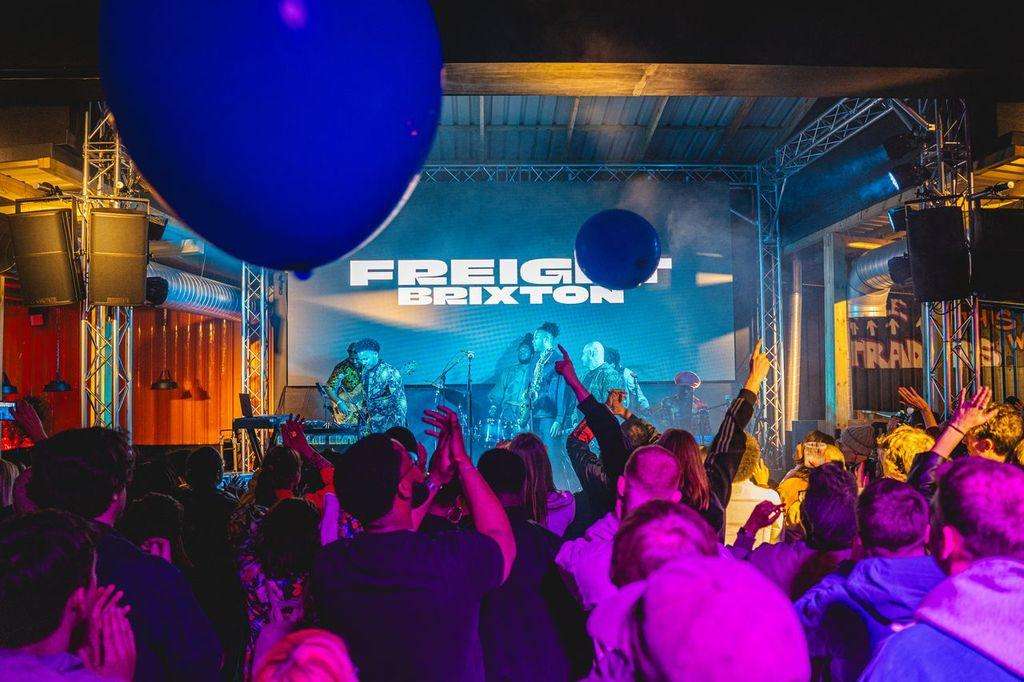 Full venue set up for a live music event with stage, band performing, large crowd, and dynamic blue and magenta lighting.